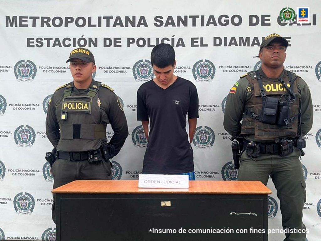 En la fotografía se aprecia a Jhonier Gaviria Segura, de pie, con los brazos atrás y la cabeza agachada. Viste camiseta negra de manga corta, pantalón negro y zapatos negros. A lado y lado de él hay uniformados de la Policía Nacional. Delante de ellos hay una mesa y sobre ella un letrero que dice: ‘Orden judicial’. Detrás de ellos hay un pendón institucional de la Policía.
