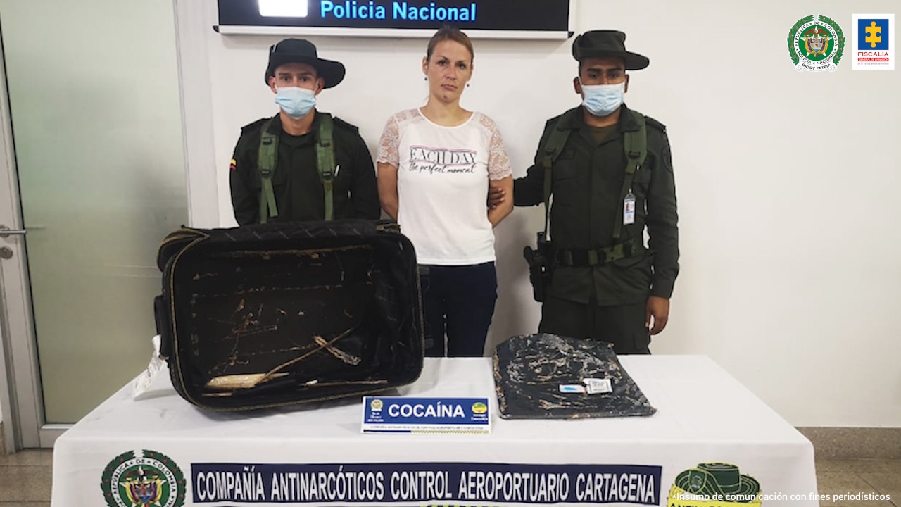 En la imagen se observa una mujer con camisa blanca y pantalón negro custodiada por dos agentes de la Policía Nacional y una mesa en la que se aprecia la maleta con la droga incautada.