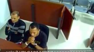 En la fotografía aparecen dos hombres sentados sobre una mesa de madera. Uno de ellos es de tez blanca, tiene los brazos cruzados y está vestido con buzo negro. El otro hombre es moreno, tiene las manos cruzadas colocadas en su mentón y está vestido con camiseta negra