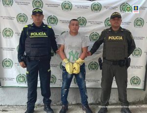 En la fotografía hay un hombre de baja estatura de pie, con los brazos esposados, vestido de camiseta gris, jean azul y tenis negros. Se encuentra custodiado por dos uniformados de la Policía Nacional.