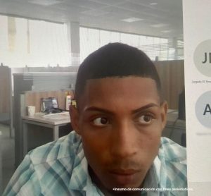 En fotografía está el rostro de un hombre delgado, moreno, ojos, nariz y boca grande, de cabellos cortos, vestido con una camisa de cuadros azules, grises y blancos. Se encuentra sentado en una silla en el interior de unas oficinas asistiendo virtualmente a la audiencia en la que se le procesó.