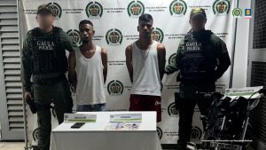  En la fotografía aparecen dos hombres delgados, morenos, de pie con los brazos atrás esposados, vestidos con camisillas blancas y bermudas. Se encuentran custodiado por dos uniformados de la Policía Nacional. 