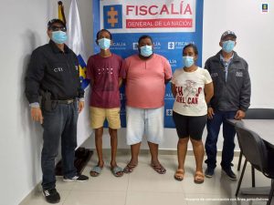 En la imagen están de pie, un hombre delgado vestido de camiseta roja con bermuda amarilla y sandalias grises, un hombre de contextura gruesa de camiseta rosada y bermuda de jean y sandalias negras y una mujer de camiseta blanca con bermuda negra y sandalia amarillas, custodiados por dos uniformados del CTI de la Fiscalía.