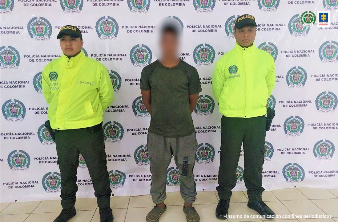 custodiado por dos hombres de la policía quienbes están uniformados, detrás de ellos hay un pendón institucionel.