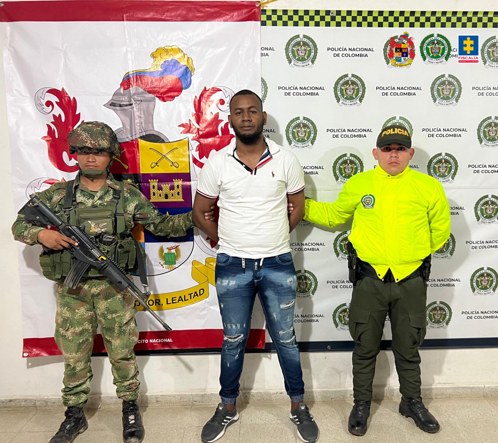 En la imagen aparece el capturado en la parte central, rodeado de dos miembros del Ejército y Policía. En la parte posterior aparecen banner de las mismas instituciones. 