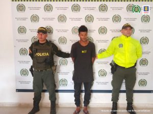 En la fotografía está el hombre condenado a 18 años de prisión por el feminicidio de su compañera sentimental, a los lados tiene 2 servidores de la Policía Nacional y atrás está el pendón de la misma institución. 