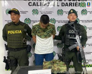 En la fotografía se aprecia de pie, con los brazos atrás y la cabeza agachada, a Jhon Stiven Ospino López. Viste camiseta de manga corta blanca y en la parte superior con estampado camuflado; jean azul. Al lado y lado de él hay uniformados de la Policía Nacional. Delante de ellos hay una mesa y sobre ella hay varios cartuchos y una chaqueta del Ejército doblada. En la parte de atrás de ellos hay un pendón institucional de la Policía Nacional.