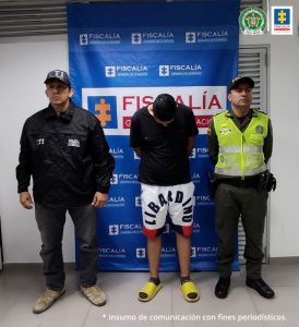 capturado de frente manos atrás, custodiado por investigador del CTI de la Fiscalía y un policía. Detrás banner de la Fiscalía.