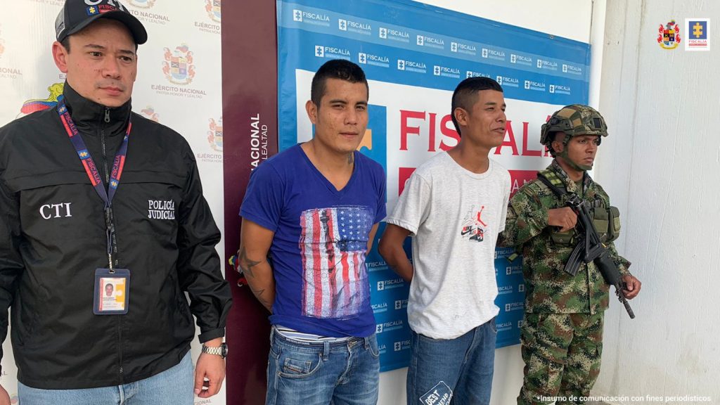 En la fotografía aparecen dos capturados junto a personal de Ejército Nacional y Fiscalía General de la Nación. En la parte posterior de la imagen se ven banners de Ejército Nacional y la Fiscalía General de la Nación. 