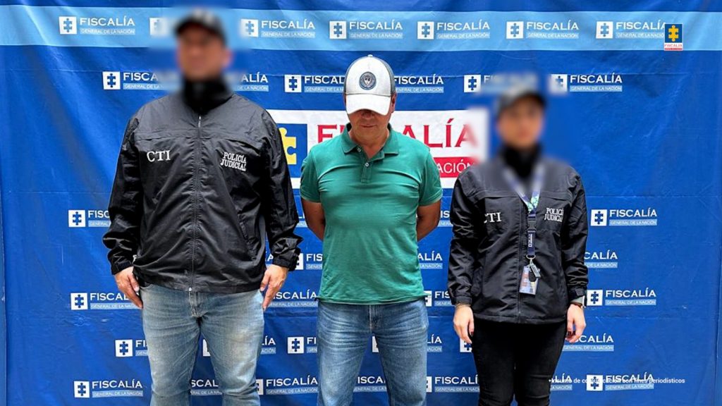 Fotografía del día de la captura del procesado usando una gorra que evita ver el rostro y dos servidores del CTI a los lados, en la parte de atrás hay un pendón azul con el logo de la Fiscalía 