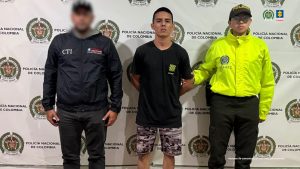  en la imagen se ve una persona detenida bajo custodia de un investigador del CTI y un uniformado de la Policía Nacional. Detrás suyo un backing institucional. 