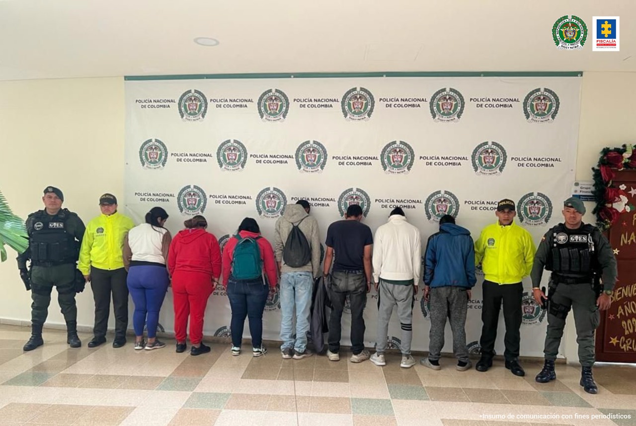 Se ven 7 capturados de espaldas y a cada lado de la fila dos funcionarios de Policía Nacional.  Atrás un pendón de Policía Nacional