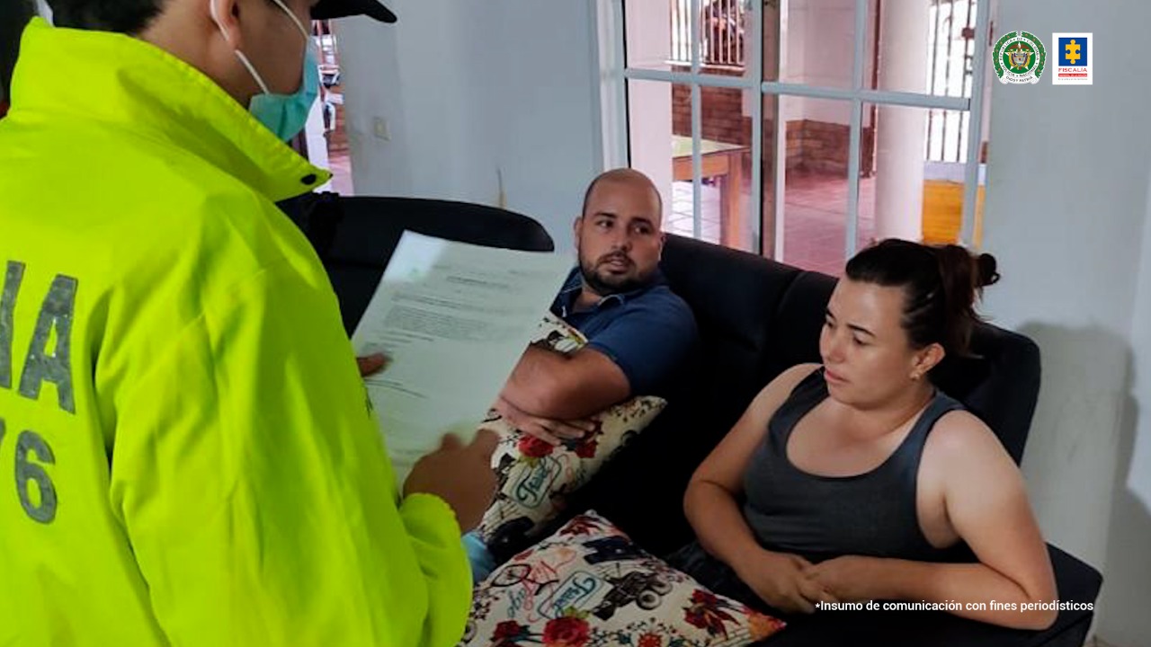En la imagen se ve a los hermanos Laura Bibiana y Víctor Daniel Claro Bonilla mientras un policía les indica cuáles son sus derechos, pues serán capturados por presuntamente incurrir en delitos de lavado de activos. Las dos personas y el uniformado al interior de una casa. 