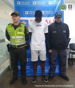 capturado de frente manos atrás, custodiado por investigador del CTI de la Fiscalía y un policía. Detrás banner de la Fiscalía