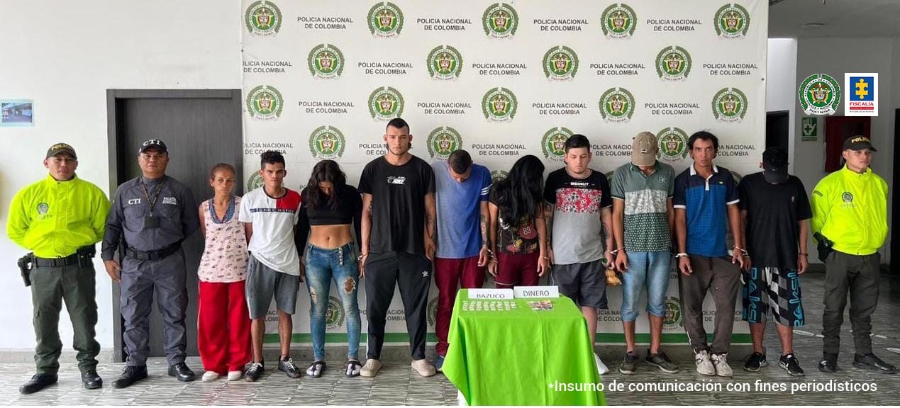 En la foto aparecen de pie y en fila los presuntos integrantes del grupo delictivo ‘Los de Papá’. Al lado derecho de ellos hay un uniformado de la Policía Nacional y un servidor del CTI de la Fiscalía. A la izquierda de ellos hay un uniformado de la Policía. Delante de ellos hay una mesa con mantel verde con varias bolsas plásticas pequeñas y dinero en efectivo, así como dos letreros que dicen ‘bazuco’ y ‘dinero’. Detrás de ellos hay un pendón institucional de la Policía. 