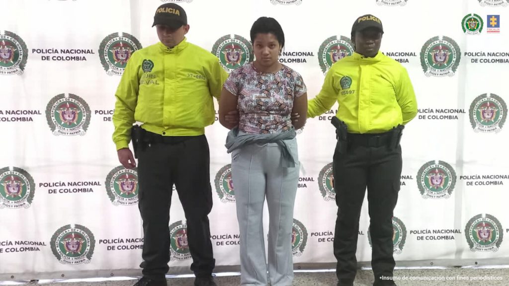 En la fotografía está la procesada con una camiseta, una sudadera gris y un saco amarrado a la cintura con dos servidores de la Policía Nacional que usan chaqueta verde fluorescente y al fondo hay un pendón de la Policía con sus logos en mosaico. 