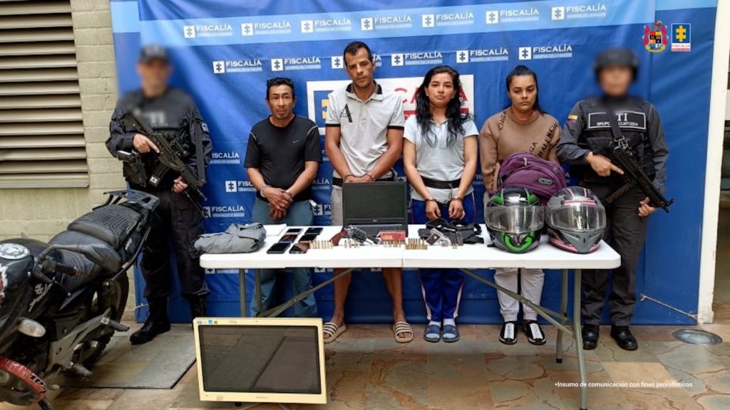 En la fotografía están las 2 mujeres y los 2 hombres que habrían participado en el homicidio con dos servidores del CTI, además, adelante hay una mesa con los computadores las armas de fuego, municiones, cascos de moto y otros elementos incautados y a un lado la motocicleta hallada en el operativo de allanamiento y registro.
