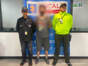 Se visualiza al capturado con el rostro difuminado junto a un investigador del CTI y un uniformado de la Policía Nacional. Detrás el banner que identifica a la Fiscalía General de la Nación 
