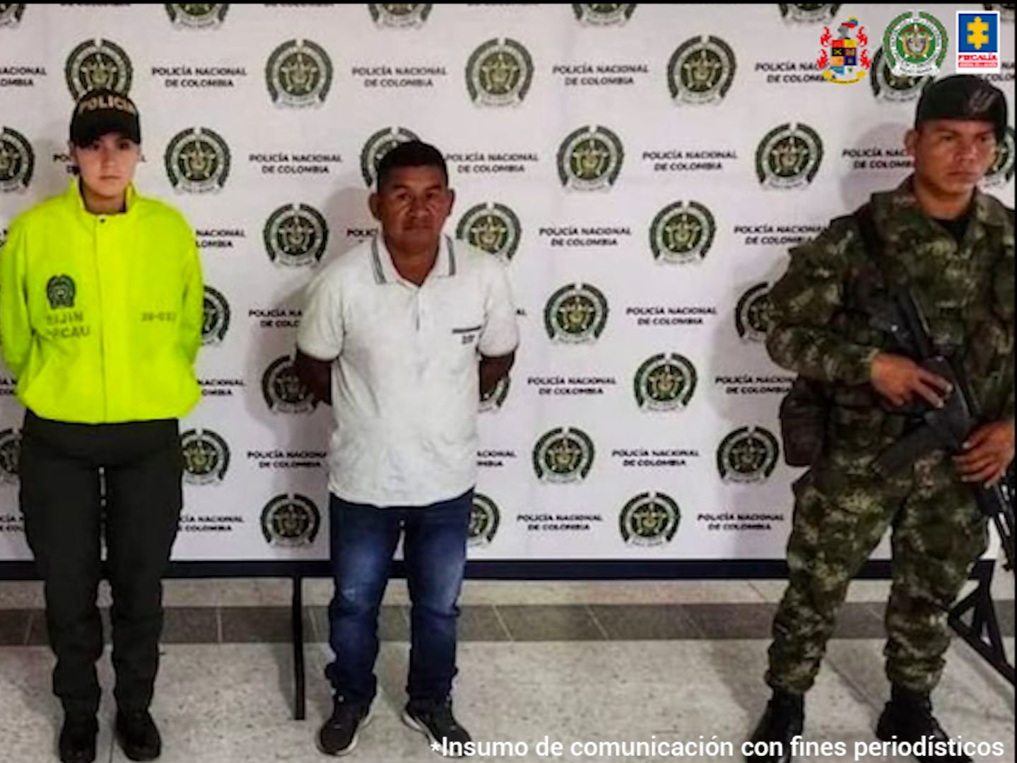 En la foto aparece Óscar Eduardo Escué Siclos, de pies, con los brazos atrás. Viste camisa blanca de manga corta, jean azul y zapatos oscuros. Al lado izquierdo de él hay una uniformada de la Policía Nacional y a su izquierda un uniformado del Ejército. Detrás de ellos hay un pendón institucional de la Policía.