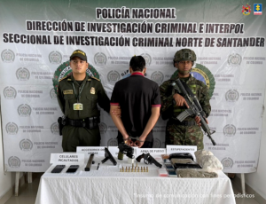 Se visualiza al procesado de espaldas junto a dos uniformados uno de la Policía Nacional y el otro del Ejército Nacional. Frente a ellos una mesa con los elementos incautados (armas, munición, sustancias estupefacientes y teléfonos celulares). Detrás de ellos el banner que identifica a la Policía Nacional