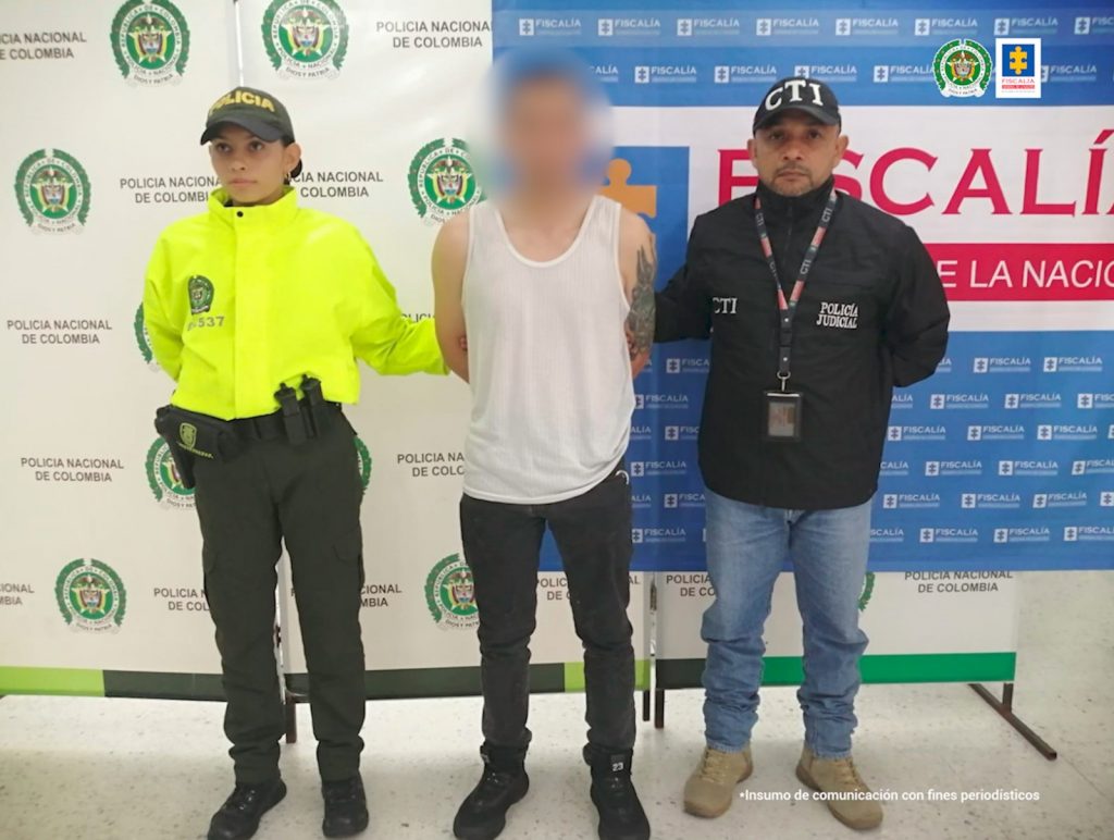  capturado de frente custodiado por un investigador CTI de la Fiscalía y un Policía. Detrás banner de Policía y de Fiscalía.