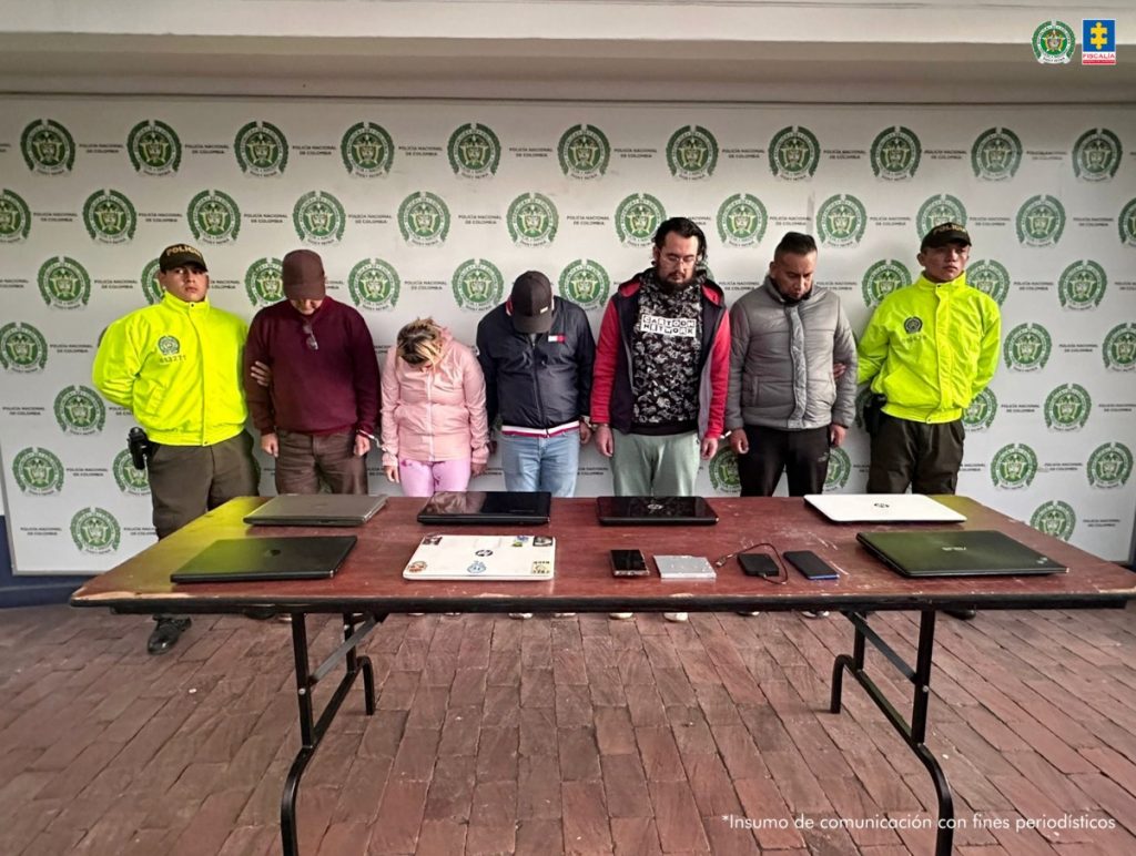 en la fotografia se puede ver a los 5 detenidos en medio de dos policias, en frente de ellos se encuentra una mesa con equipos con los cuales realizaban los delitos. Atras de todos baking de la policia