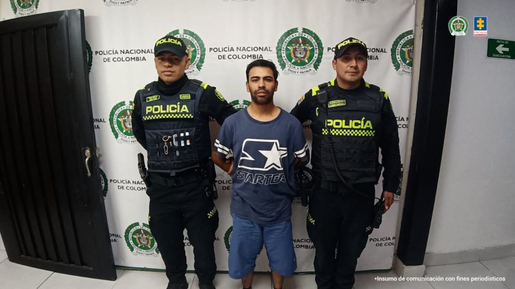 Fotografia en donde se ve al capturado custodiado por dos policias, dentras de ellos se ve un pedon de la policia nacional