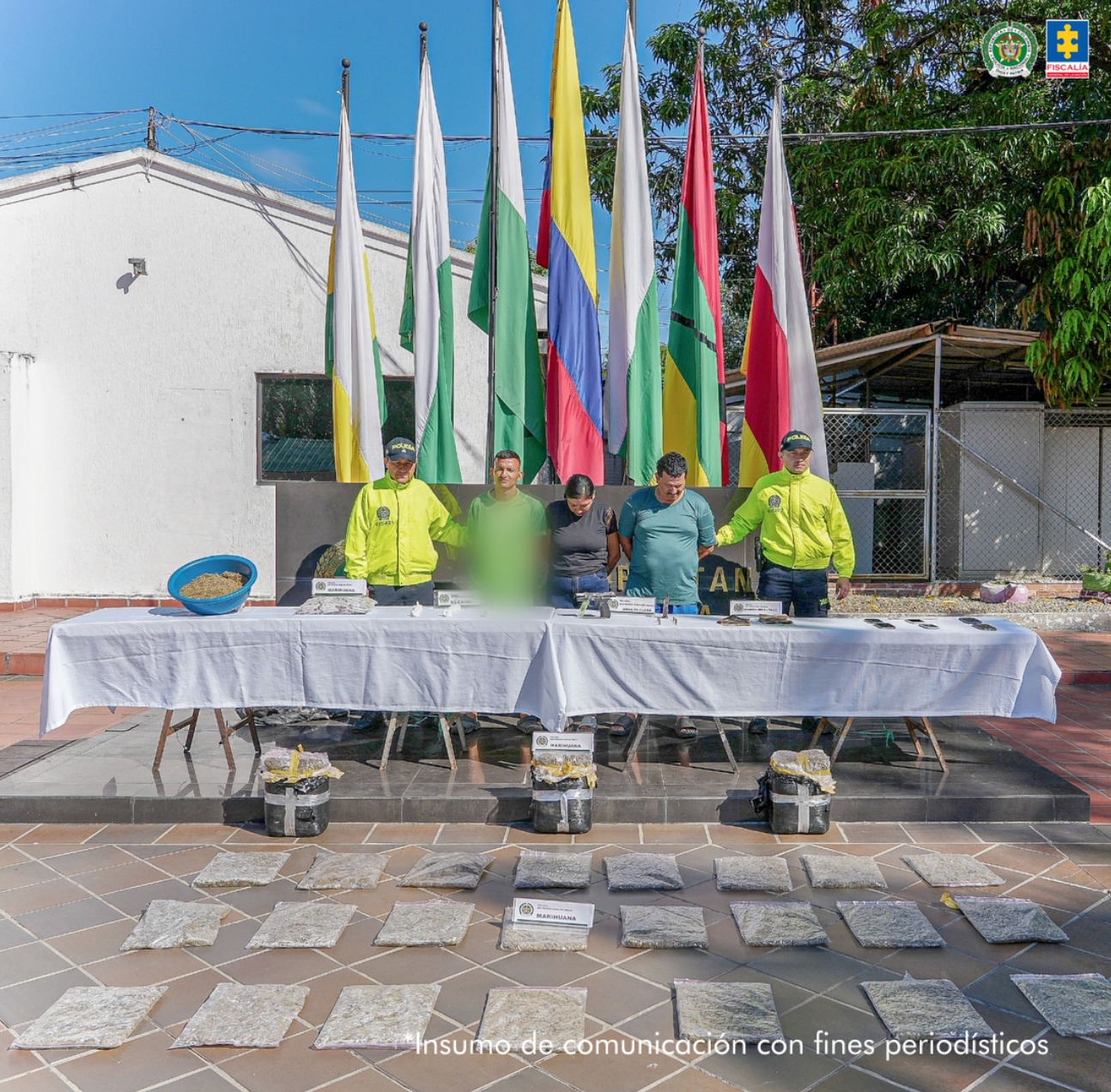 Los capturados se encuentran de frente y custodiados por dos uniformados de la Policía Nacional. En la foto se ve de fondo unas banderas, una mesa con el material incautado. En el piso se observa el estupefaciente incautado durante los registros de allanamientos.