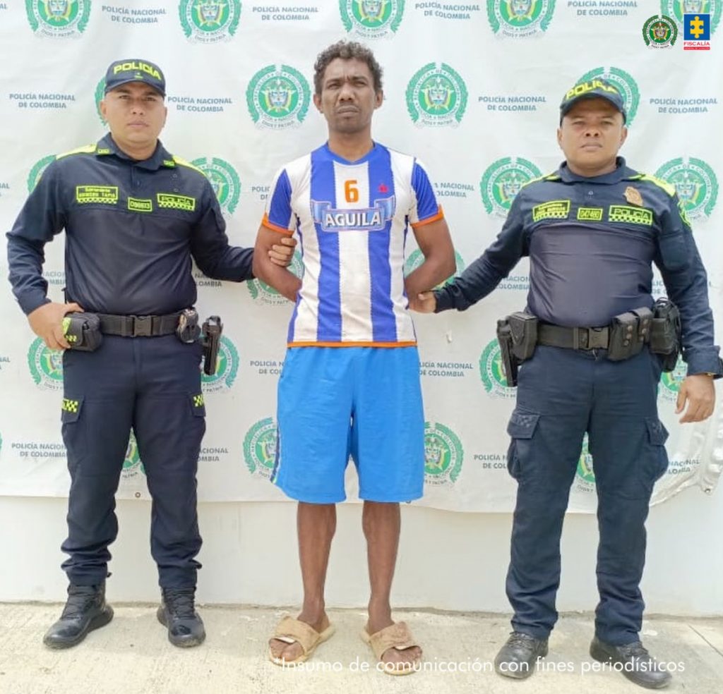 En la imagen aparece una persona con camiseta deportiva, de rayas blancas y azules, entre dos uniformados de la Policía Nacional. 