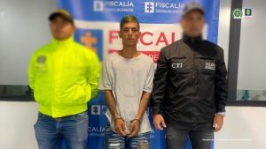 Se visualiza al procesado junto a un uniformado de la Policía Nacional y un investigador del CTI. Detrás el banner que identifica a la Fiscalía General de la Nación.