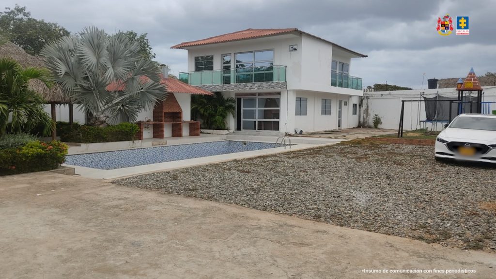 La imagen muestra una lujosa propiedad de dos pisos ubicada en un terreno amplio. La casa es blanca, con techo de tejas rojas y balcones con barandas de vidrio. Frente a la casa hay una piscina vacía y al lado de esta, una zona de asador en ladrillo rojo con techo de palma. También hay un jardín con plantas ornamentales, un parque infantil con un pequeño castillo de juegos, una cama elástica y un carro blanco estacionado cerca. El cielo está nublado y el lugar parece estar en una zona tranquila. En la esquina superior derecha aparecen los logos de la Fiscalía General de la Nación y otro escudo institucional. En la parte inferior derecha, hay un pequeño texto que dice: "Insumo de comunicación con fines periodísticos".