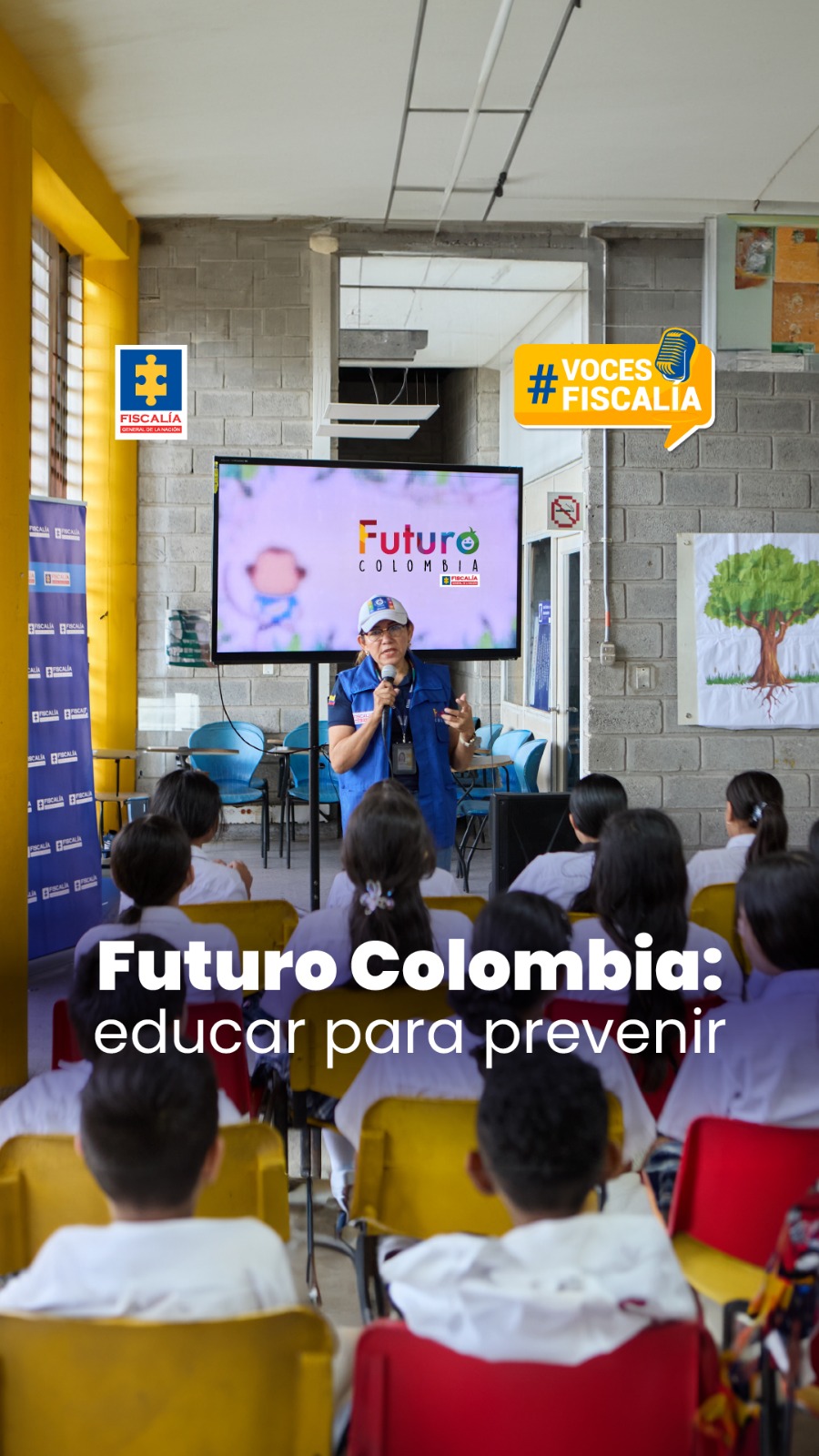 La imagen muestra una jornada educativa liderada por la Fiscalía General de la Nación en el marco del programa "Futuro Colombia", cuyo lema es “educar para prevenir”. En la fotografía se observa a una funcionaria con micrófono, chaleco azul institucional y gorra, hablando frente a un grupo de estudiantes que están sentados y atentos a la charla. Detrás de ella hay una pantalla grande con el logo del programa y el nombre de la Fiscalía. El entorno es un aula o salón amplio, con paredes de bloque gris, sillas de colores (amarillas, rojas y azules), y carteles institucionales. Se destaca un ambiente educativo y participativo. En la parte superior de la imagen aparecen los logotipos de la Fiscalía y el sello gráfico de la campaña “#VocesFiscalía”,