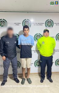 En la imagen se observa a un hombre joven, de pie en el centro, con las manos hacia atrás, vestido con una camiseta azul clara y negra, pantalón corto beige y sandalias oscuras. A su lado izquierdo está un funcionario del CTI con chaqueta negra y gorra, cuyo rostro aparece difuminado. A la derecha, un uniformado de la Policía Nacional viste chaqueta amarilla fluorescente y gorra negra. Al fondo se aprecia un muro blanco con los logos de la Policía Nacional de Colombia y de la Fiscalía General de la Nación.
