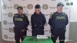 El capturado aparece al centro y en cada costado un uniformado de la Policía Nacional. Atrás un pendón de Policía Nacional y al frente una mesa con un arma y municiones incautadas.