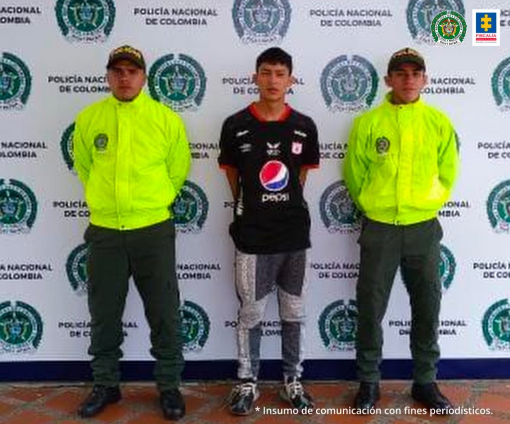 Se observa el capturado en el centro y de frente. En cada constado un funcionario de Policía Nacional. Al fondo se ve pendón de Policía Nacional