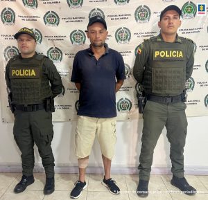 En la imagen se ve una persona detenida bajo custodia de dos integrantes de la Policía Nacional. Detrás suyo un backing institucional.   