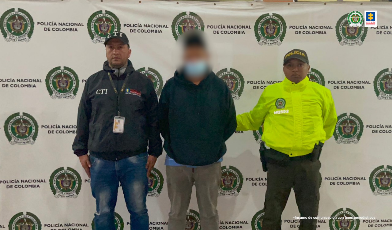 En la imagen se observa a un hombre que es custodiado por un policía y un integrante del CTI. La persona capturada sería responsable del asesinato de su hijastro. Al fondo se observa un cartel con logos de la Policía Nacional.  
