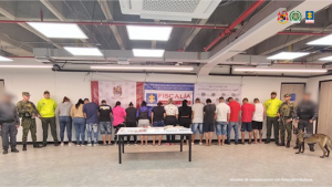 Se observa a los procesados de espaldas junto a investigadores del CTI y uniformados del Ejército y Policía Nacional. Frente a ellos una mesa con los elementos incautados. Detrás de ellos los banners institucionales de la Fiscalía General de la Nación y de Ejército y Policía Nacional.