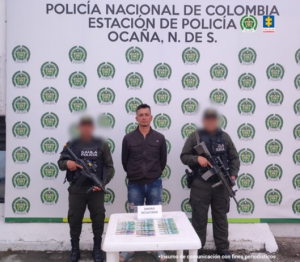  Se visualiza al capturado junto a dos uniformados de la Policía Nacional. Frente a ellos en una mesa el dinero exigido. Detrás de ellos el banner que identifica a la Policía Nacional.