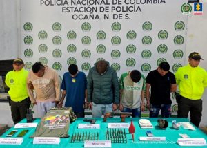 Se observan a cinco de los capturados junto a dos uniformados de la Policía Nacional. Frente a ellos en una mesa los elementos incautados: una escopeta, un arma traumática, munición, sustancias estupefacientes, teléfonos celulares, grameras, inhibidor de señal, pólvora y prenda alusiva al frente 41 de las Farc. 