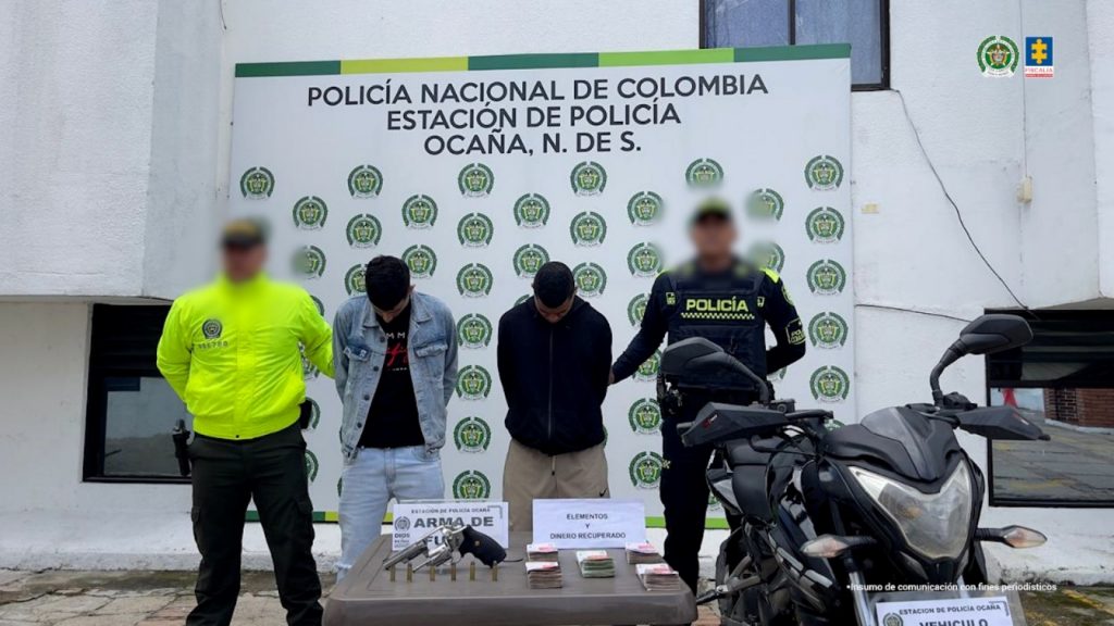 Se visualiza a los dos capturados junto a dos uniformados de la Policía Nacional. Frente a ellos una mesa con el dinero recuperado y los elementos incautados utilizados para el hurto un arma de fuego y la motocicleta. Detrás de ellos el banner que identifica a la Policía Nacional en Ocaña.