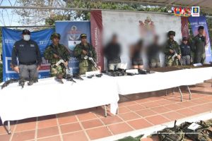 Se visualiza a los tres capturados con los rostros difuminados junto a dos investigadores del CTI y uniformados del Ejército y Armada Nacional. Frente a ellos una mesa con el material de guerra incautado (fusiles, munición y prendas). En la parte posterior los banners institucionales de la Fiscalía General de la Nación y del Ejército y Armada Nacional.