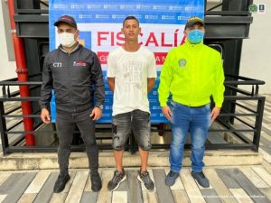 Se visualiza al capturado junto a un investigador del CTI y uno de la Sijín.  Al respaldo el banner que identifica a la Fiscalía General de la Nación.
