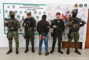  En la imagen se visualiza al capturado de espaldas junto a uniformados del Gaula de la Policía y Ejército Nacional.  En la parte posterior se observan los banners que identifican a la Policía y Ejército Nacional.