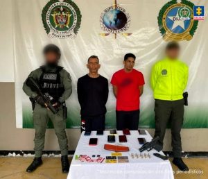 Se visualiza a los dos capturados junto a uniformados de la Policía Nacional. Frente a ellos una mesa con los elementos incautados como arma, munición y teléfonos celulares. Detrás de ellos un banner que identifica a la Policía Nacional.