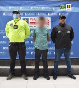 Se visualiza al capturado junto a un uniformado de la Policía Nacional y un investigador del CTI. Detrás de ellos el banner que identifica a la Fiscalía General de la Nación.
