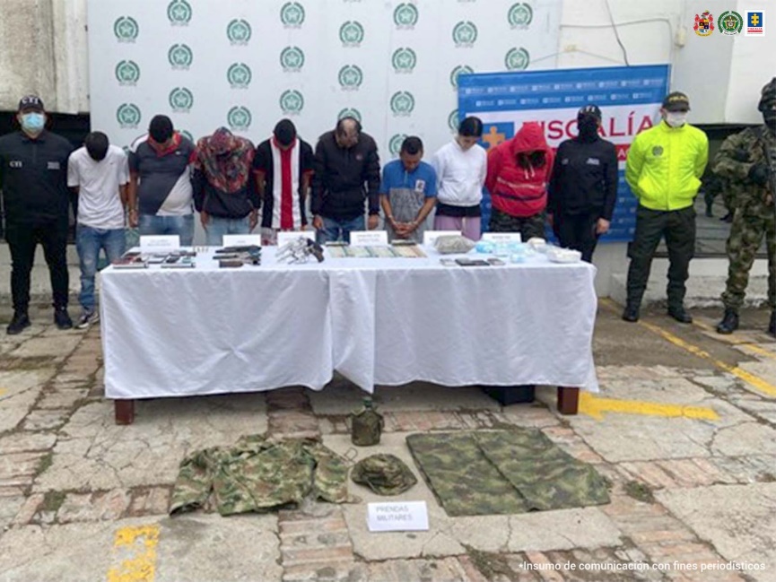 En la fotografía se aprecia los 10 detenidos junto a personal del CTI y uniformados de la Policía y del Ejército Nacional. Frente a ellos se ubica una mesa con los elementos materiales incautados como armas, sustancias estupefacientes, dinero y material de intendencia. En la parte posterior los banners que identifican a la Policía Nacional y Fiscalía General de la Nación