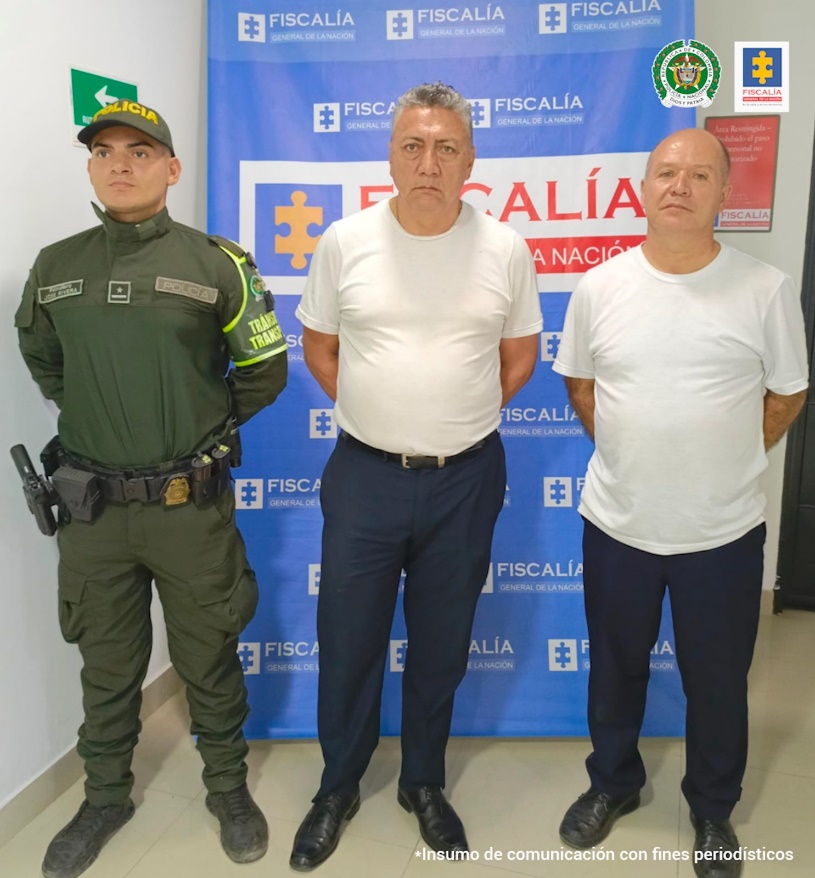 Se visualiza a los dos capturados junto a un uniformado de la Policía Nacional. Detrás se encuentra el banner de la Fiscalía General de la Nación