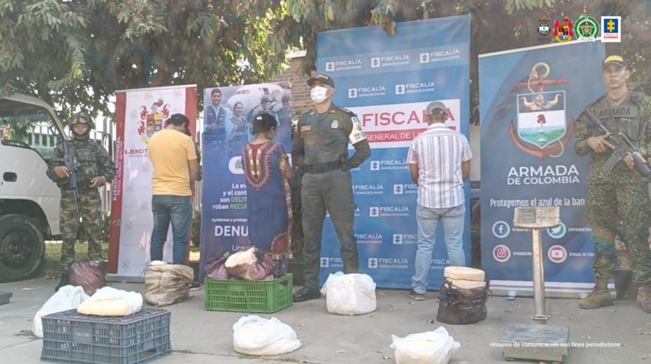 Se observa a los procesados junto a uniformados de la Policía y Ejército Nacional. Frente a ellos el material incautado. En la parte posterior los banners institucionales que identifican a la Fiscalía General de la Nación y a la Policía, Ejército y Armada Nacional.