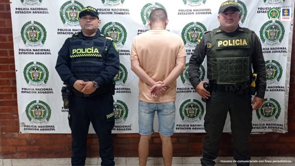 En la imagen se observa al capturado de espalda junto a dos uniformados de la Policía Nacional. Detrás de ellos se visualiza el banner que identifica a la Policía Nacional de Aguazul (Casanare) 